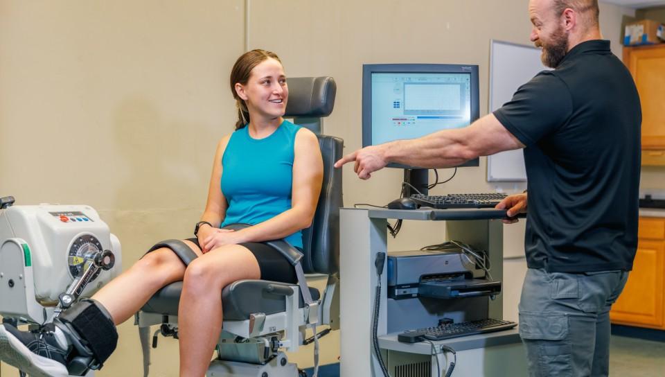 A student sitting in a machine that measures muscle strength as their professor shows them the results on a monitor